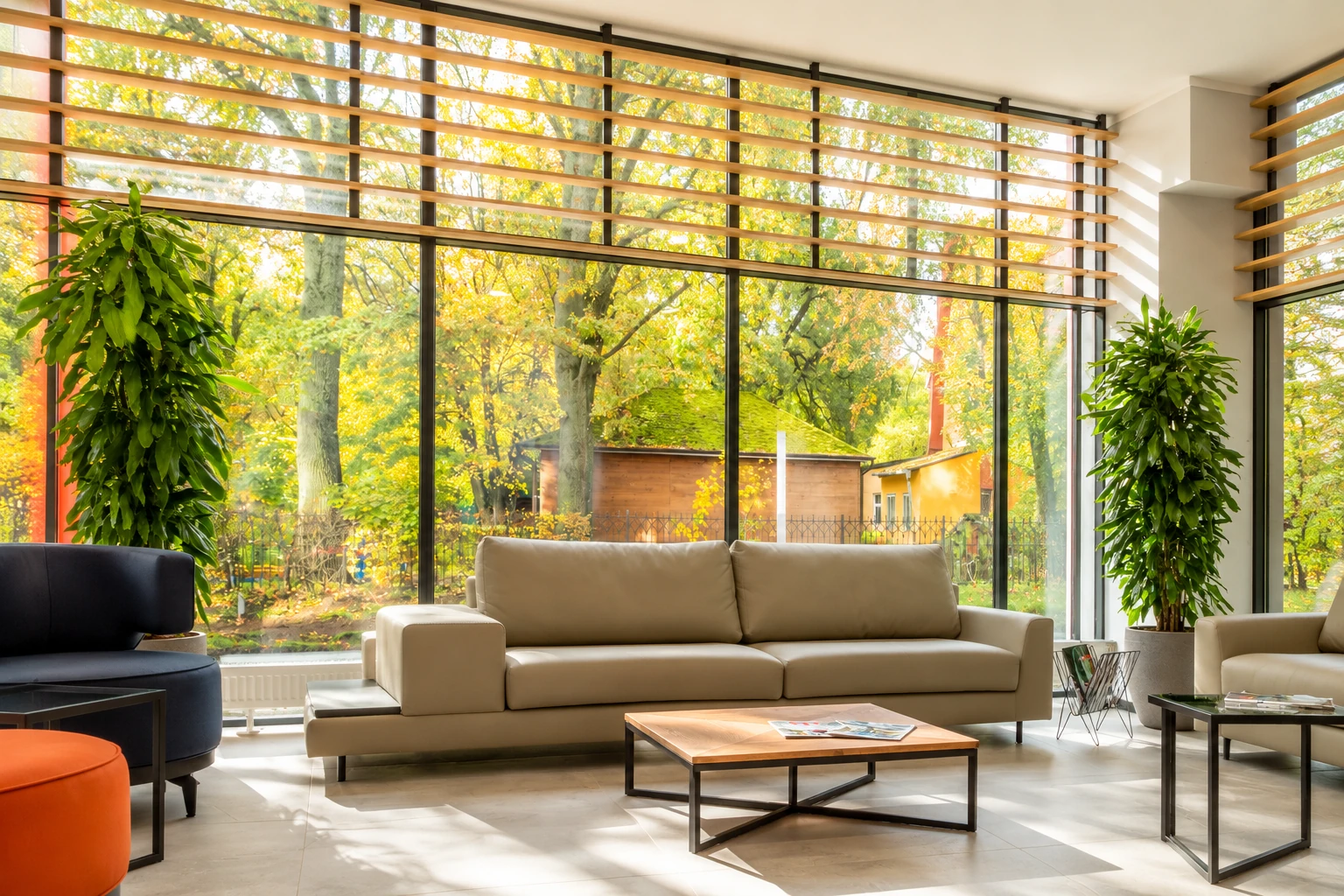 Bright living room with a tan sofa and floor-to-ceiling windows overlooking autumn trees.