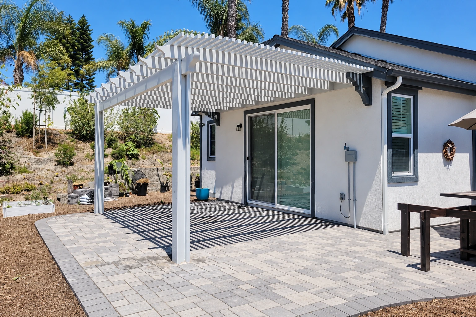 White slatted patio cover over a paved patio beside a house, casting striped shadows.
