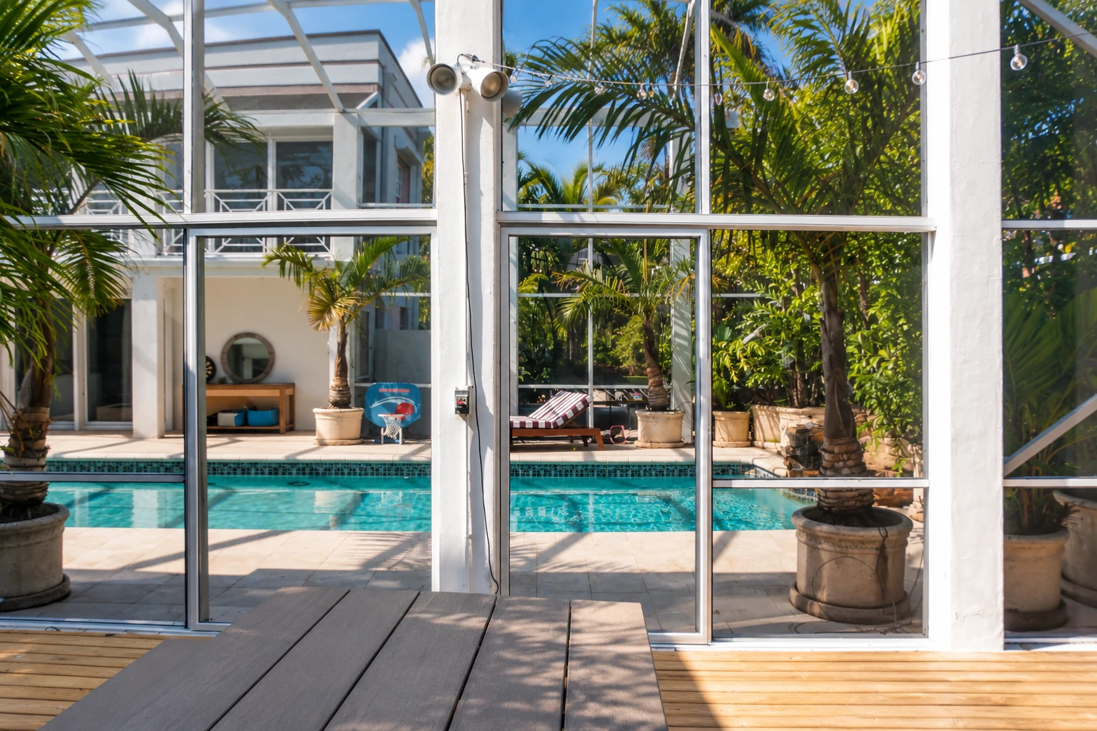Patio enclosure with white framing and glass panels around a swimming pool, with palm trees and string lights beyond.