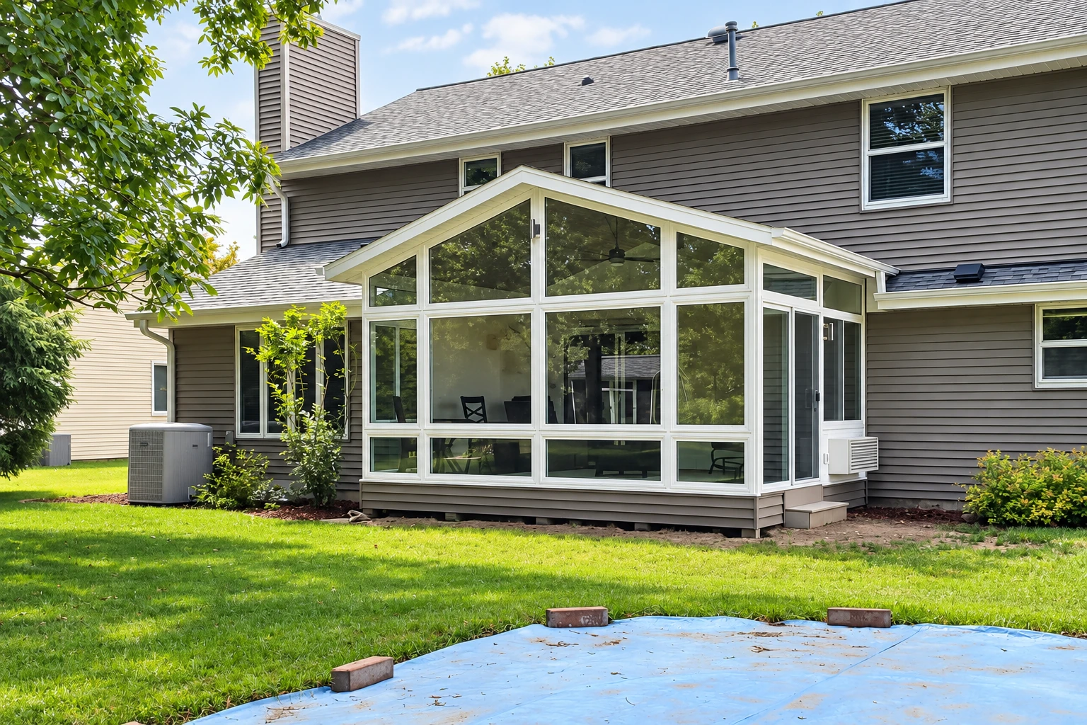 White-framed, peaked glass sunroom addition on a gray-sided house, with a lawn and patio in the foreground.