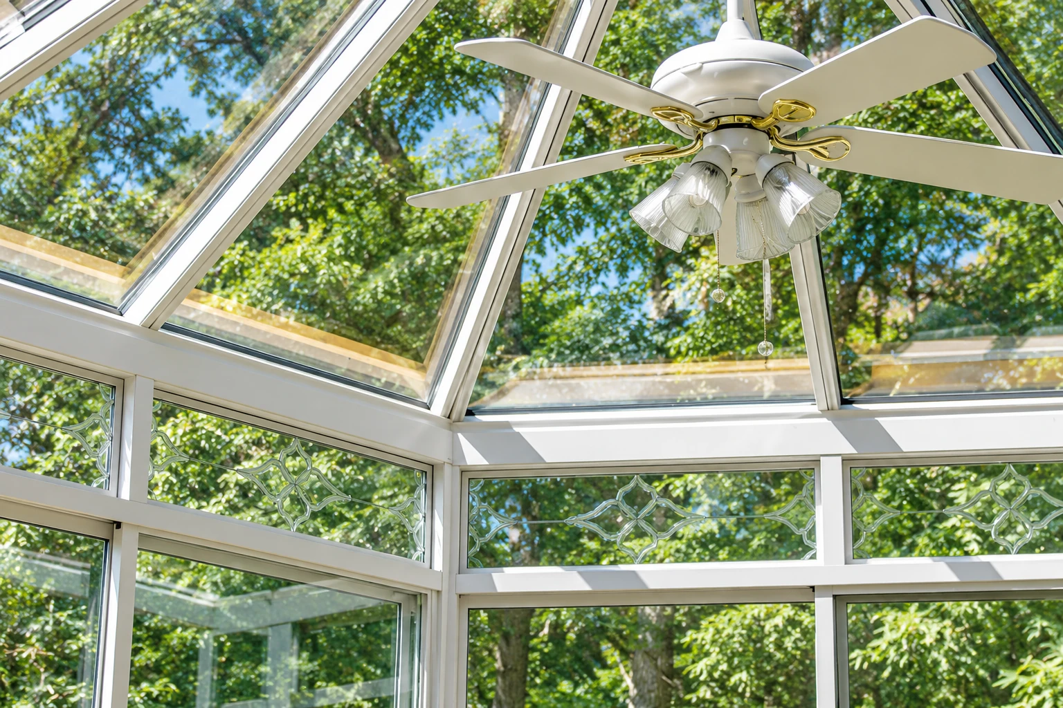 White-framed solarium ceiling with large glass panels and a central ceiling fan beneath leafy trees.