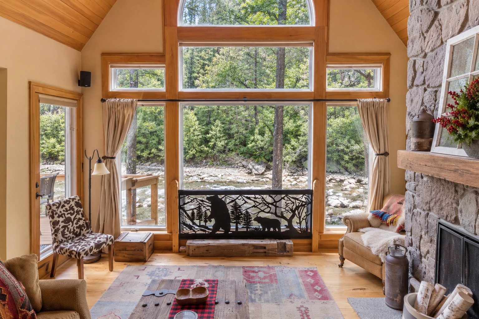 Sunroom addition with vaulted wood ceiling, tall windows overlooking a rocky stream, and a stone fireplace.