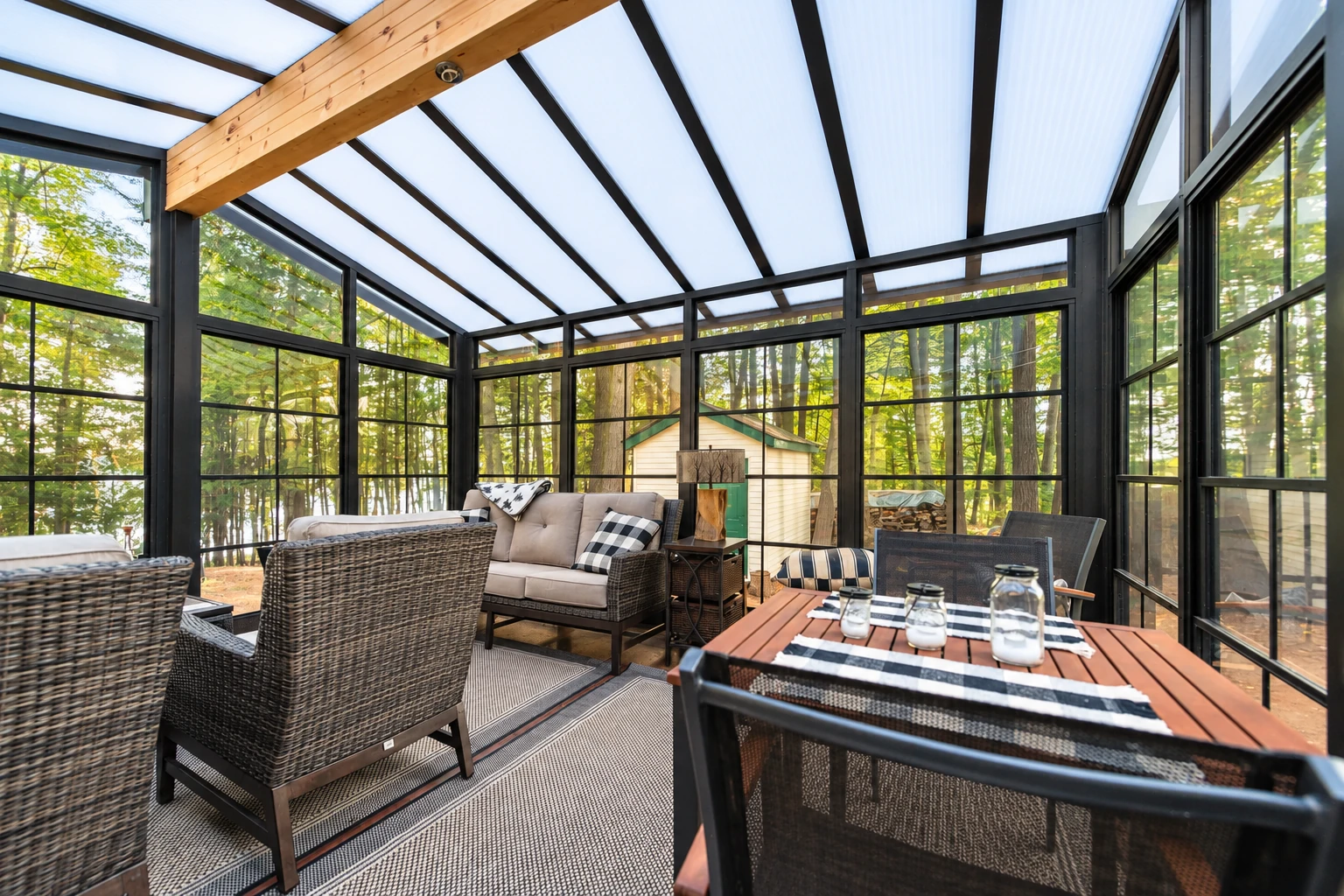 Furnished sunroom with black-framed windows, a translucent pitched roof, wicker seating and a dining table overlooking woods.
